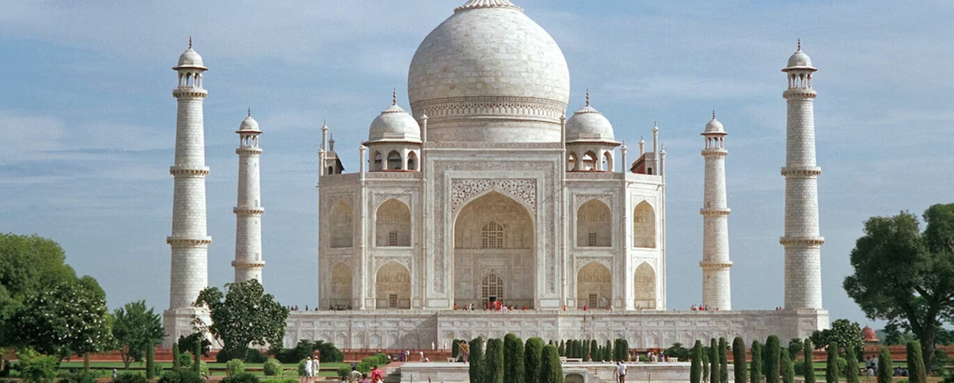 A stunning white marble mausoleum, the Taj Mahal in India, standing tall against a blue sky with intricate architectural details and reflecting in a serene pool of water. Travellers may visit this location after having their vaccinations.